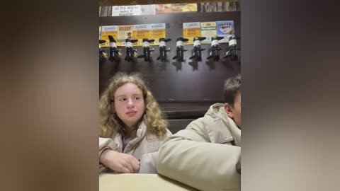 Media: Video of a young girl with curly blonde hair, wearing a beige coat, sitting in a fast-food restaurant. Behind her, a row of black figurines in police uniforms and a menu board are visible.