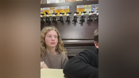 Media: Video of a young girl with curly hair, wearing a beige sweater, seated at a table in a dimly lit restaurant. Behind her, black and white toy figures hang from the ceiling.