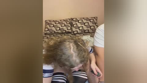 Media: Video of a blonde woman in a white and navy striped top, kneeling on a patterned cushioned bench, with a man's legs visible in the background.