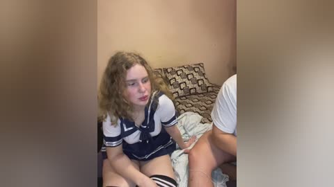 Media: A video of a young woman with curly hair in a navy sailor dress, sitting on a bed with a patterned pillow, holding her crotch.