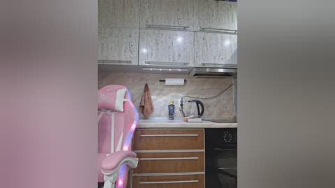 Media: A video of a small, modern kitchen with white cabinets, a pink gaming chair, wooden drawers, a beige countertop, a paper towel holder, and a kettle.