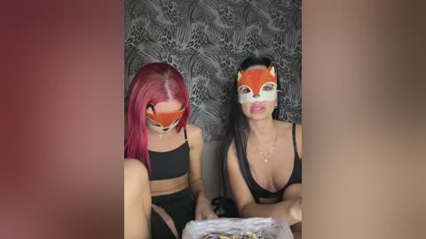Video of two women in black lingerie with red fox masks; one has bright red hair, the other black hair. They sit on a bed with leopard-print wall behind.