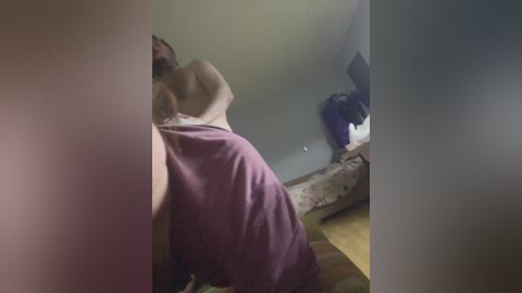 Video of a nude man with a muscular build, short brown hair, and a light skin tone, lying on a bed with purple sheets. The room has a cluttered, dimly lit atmosphere with a TV and clothes visible in the background.