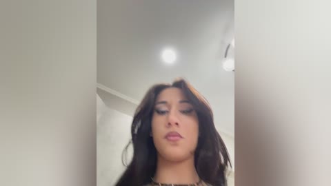 A video of a young woman with long, dark hair and light skin, wearing makeup, standing in a brightly lit room with white walls and a ceiling light.