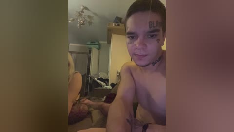 Video of a young woman with light skin and dark hair, partially nude, sitting on a bed in a dimly lit bedroom. She has tattoos on her arm and face.