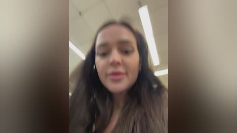 A blurry, close-up video of a young woman with long brown hair, wearing a dark top, taken indoors with fluorescent lighting.