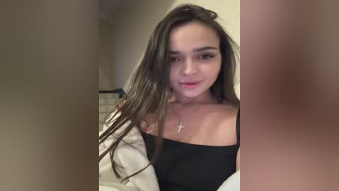 Video of a young woman with long, straight brown hair, fair skin, and light makeup, wearing a black off-shoulder top and a cross necklace. She stands in a beige hallway with blurred background.
