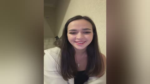Video of a smiling young woman with long dark hair, wearing a white hoodie and black top, standing in a dimly lit hallway with beige walls and tiled floor.