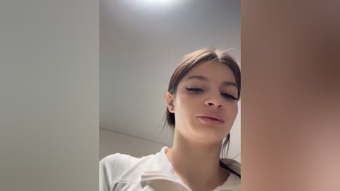 Video of a young woman with light skin, brown hair, and closed eyes, leaning against a beige wall in a dimly lit room. She wears a white shirt.