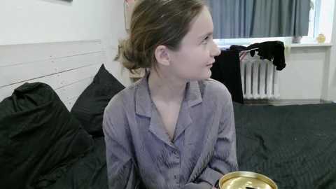 Video of a young woman with light skin and brown hair tied back, wearing a purple button-up pajama shirt, seated on a bed with black sheets and pillows, holding a golden cup, in a dimly lit, minimalistic bedroom.