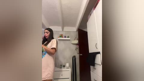 Video of a woman in a beige T-shirt, standing in a small, dimly-lit bathroom with white walls and shelves. She appears to be talking on a phone.