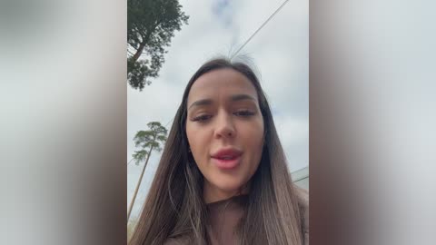 A video of a young woman with long brown hair, fair skin, and a light brown top, smiling against a cloudy sky with palm trees in the background.