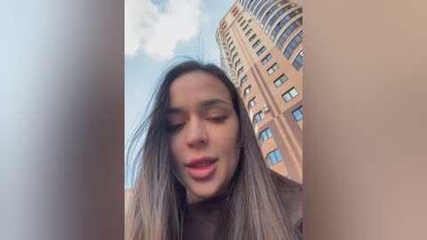 A video of a young woman with long, straight brown hair, wearing a light-colored top, smiling, taken from a low angle, with a tall, beige, windowed building in the background under a partly cloudy sky.