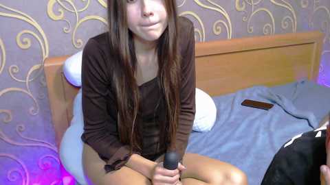 A video of a young woman with long brown hair, sitting on a bed in a room with patterned wallpaper, holding a microphone. She wears a brown top and beige shorts.