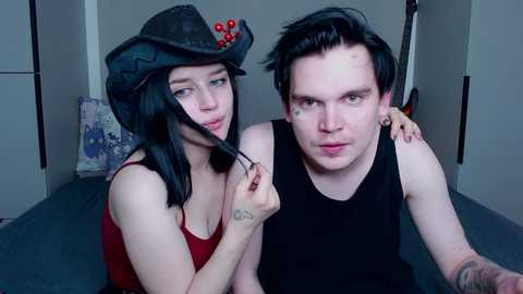 Video of a pale-skinned, tattooed woman with black hair and a cowboy hat, holding a feather, and a pale-skinned, tattooed man in a black tank top, both with serious expressions.