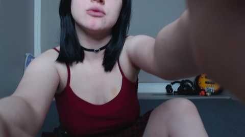Video of a young woman with shoulder-length black hair, fair skin, and a red tank top, sitting on a gray couch. She has a black choker and a visible tattoo on her arm. Background includes a toy car and a door.