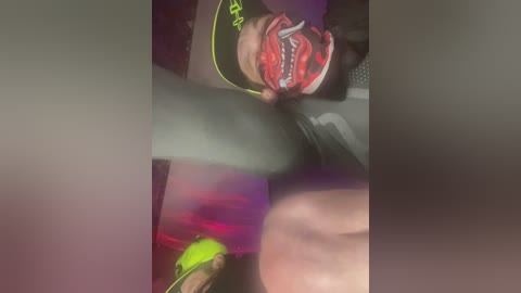 A blurred video of a person lying on their back, with a neon green object and red object visible. The background is dark, making the objects stand out.