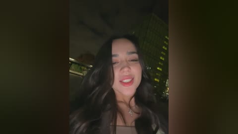 A video of a smiling woman with long, dark hair, wearing a light-colored top, taken indoors with a dimly lit background featuring blurred green foliage.