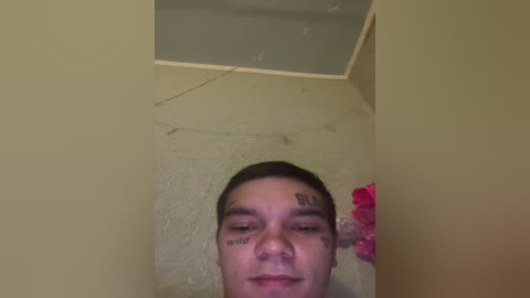 A video of a young man with short dark hair, wearing a black t-shirt, lying on a beige carpeted floor with a pink object nearby. His face is covered with black tattoos.