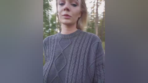 Video of a Caucasian woman with fair skin and light brown hair, wearing a gray cable-knit sweater, standing in a forest with tall pine trees in the background.