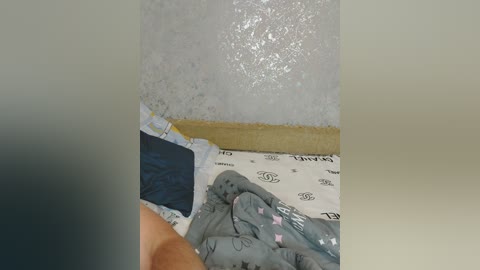 Video of a person's bare leg on a bed with a grey quilt, white sheet, and a pillow with a blue pillowcase. The wall behind is covered in a textured, white, textured wallpaper.