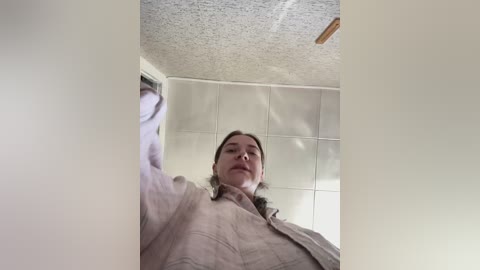 Video of a woman with light skin and brown hair, wearing a beige jacket, standing in a room with white tiled walls and a textured ceiling.