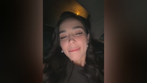 Video of a woman with long black hair, fair skin, and closed eyes, smiling softly, wearing a black shirt, in a dimly lit room with a warm light source in the background.