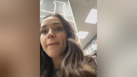 Video of a young woman with long dark hair, smiling, in a modern office with white walls and fluorescent lights.