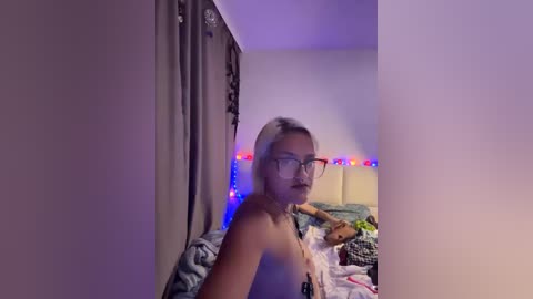 A video of a topless woman with light skin and short, platinum blonde hair, wearing glasses and a necklace, standing in a dimly lit bedroom with a stuffed teddy bear, a bed, and colorful string lights.