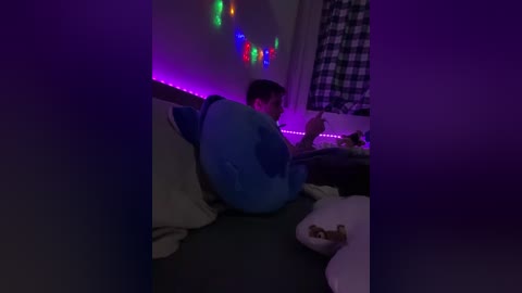 Video of a young man in a blue hoodie and dark pants sitting on a bed with a checkered wall and colorful string lights in the background.