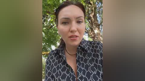 A video of a fair-skinned woman with brown hair, wearing a black patterned blouse, standing outdoors among greenery. She appears serious, with natural lighting.