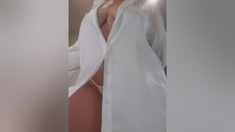 Video of a partially undressed woman in an oversized white button-down shirt, revealing her medium-sized breasts and a glimpse of white underwear. Background is blurred, emphasizing her form.