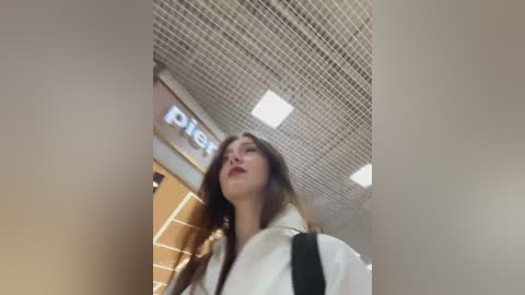 Video of a young woman with long brown hair, wearing a white jacket, standing in a modern indoor space with grid-tiled ceiling and fluorescent lights.