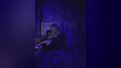 A dimly lit video shows a woman in a black dress and a man in a dark shirt, cuddling on a bed with a patterned blanket, under a purple-hued light.