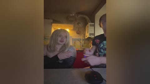 Video of a dimly lit room with a blonde woman wearing a black top and a man in a green plaid shirt, both holding a controller.