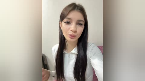 Video of a young woman with long, straight dark hair, fair skin, and light makeup. She wears a white shirt and pouts slightly, standing in front of a plain beige wall.