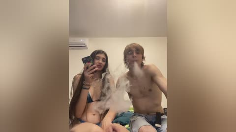 Video of two shirtless young people, one with long hair, the other with short hair, exhaling smoke in a dimly lit room.