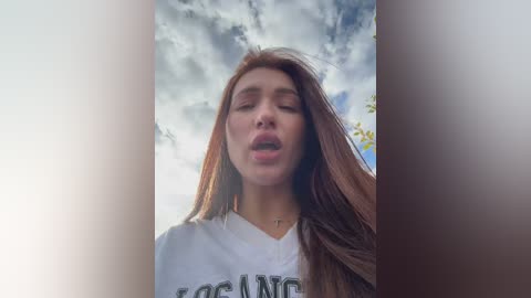 Video of a young woman with long brown hair, wearing a white T-shirt, smiling broadly against a bright, cloudy sky background.