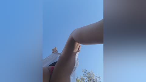 Video of a woman with light skin, wearing a white tank top and red bikini bottoms, bending over against a clear blue sky.