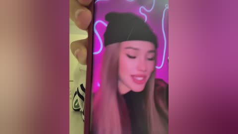 Video of a smiling young woman with long, straight brown hair, wearing a black beanie and a black top, against a pink background with neon light outlines.