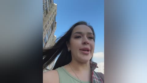 Video of a young woman with long dark hair, light skin, and wearing a green top, standing in front of a modern building against a clear blue sky.
