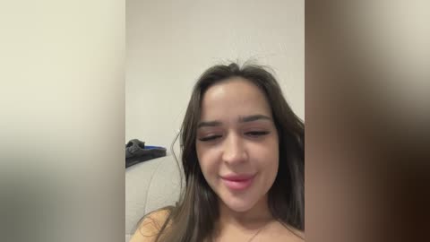 Video of a young woman with medium brown skin, dark hair, and a slight smile, standing in a white-walled room with a gray towel hanging on a rack in the background.