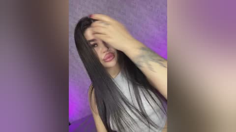 Video of a young woman with long, straight black hair, wearing a sleeveless gray top, covering her face with her left hand, against a textured, purple wall.