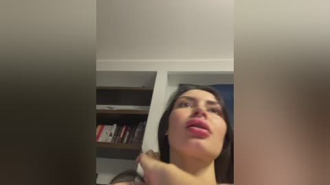 Video of a young woman with fair skin and dark hair, slightly distorted, looking up at a ceiling. She is holding a large white object. Background shows a bookshelf with books and a partially visible blue wall.