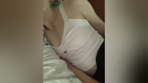 A video of a light-skinned person with green hair, wearing a white tank top and black shorts, lying on a bed with a white and green striped pillow.