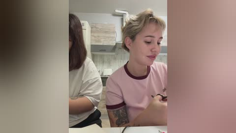 Video of two young women in a modern kitchen: one with short blonde hair and tattoos, wearing a pink shirt, focused on her phone; the other with long brown hair, dressed in a white shirt, turned away.
