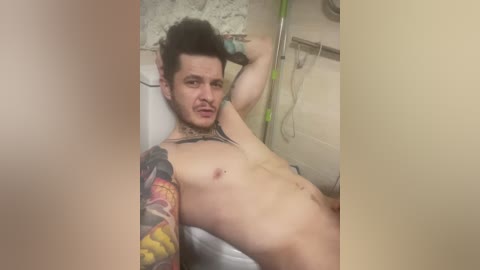 A video of a shirtless, tattooed man with dark hair lying on a white bed in a dimly lit room. The background shows a beige wall and a green medical device.