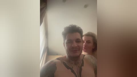 A video captures two tattooed individuals, a man with a muscular build and a woman, both smiling, in a dimly lit room with soft lighting.