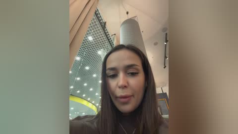 A video of a young woman with long, straight brown hair, wearing a brown turtleneck, standing indoors near a modern ceiling with a perforated metal pattern and cylindrical light fixtures.
