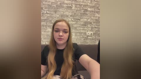 Video of a young woman with long, light brown hair, wearing a black T-shirt, sitting on a couch in front of a brick-patterned wallpaper.
